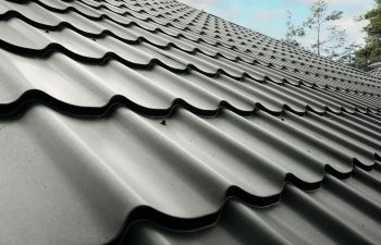 Close-up of a metal roof with overlapping, wavy panels. The roof surface is dark with a slight sheen, and trees are visible in the background under a partly cloudy sky.